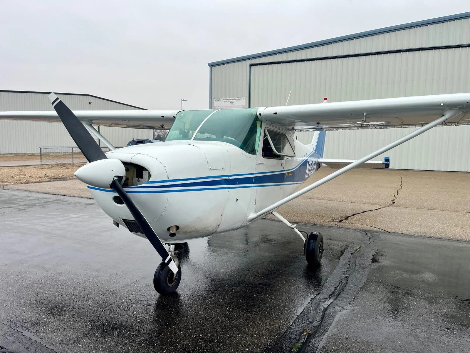 A front view of a white Cessna 172 Skyhawk with blue stripes, registration N7820X, parked on a wet tarmac.