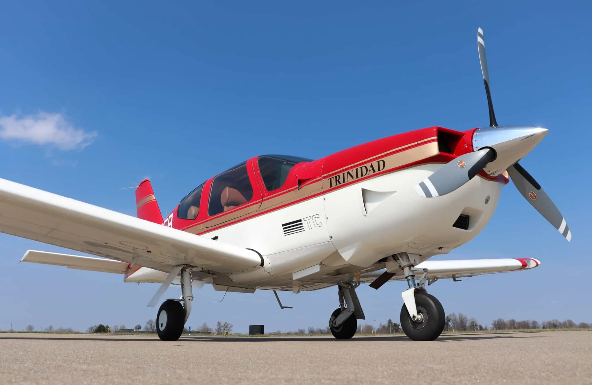 A low-angle view of a red and white Socata Trinidad aircraft with gold stripes parked on an airport tarmac.