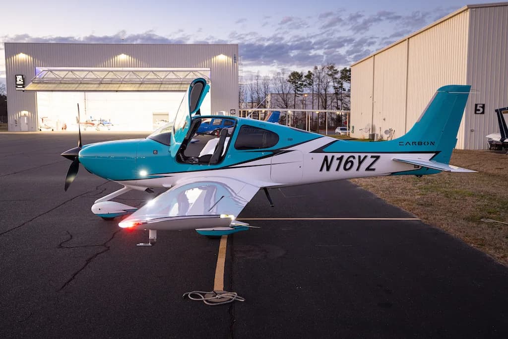 A turquoise and white Cirrus SR22 aircraft with its doors open, parked on an airfield in front of hangars at dusk.