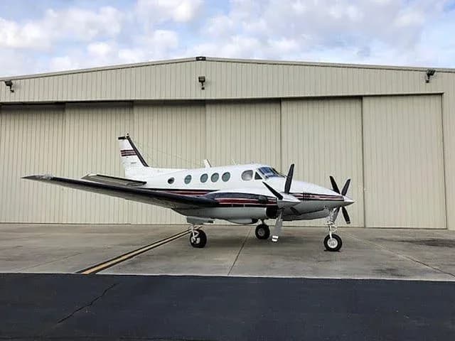 A white Beechcraft King Air twin-turboprop aircraft with dark red and black stripes is parked on an asphalt ramp in front of a light-colored hangar.