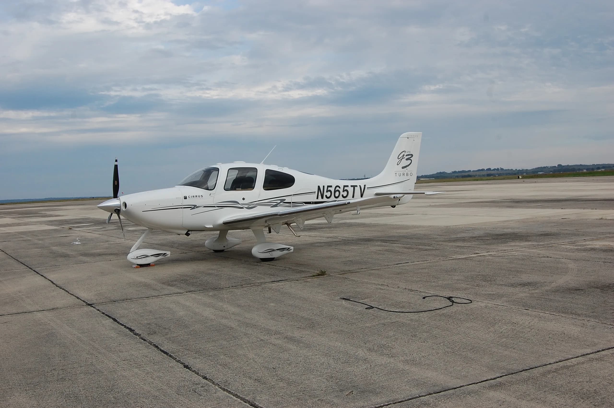A white Cirrus SR22T aircraft, N565TV, parked on a concrete tarmac with a rope on the ground.