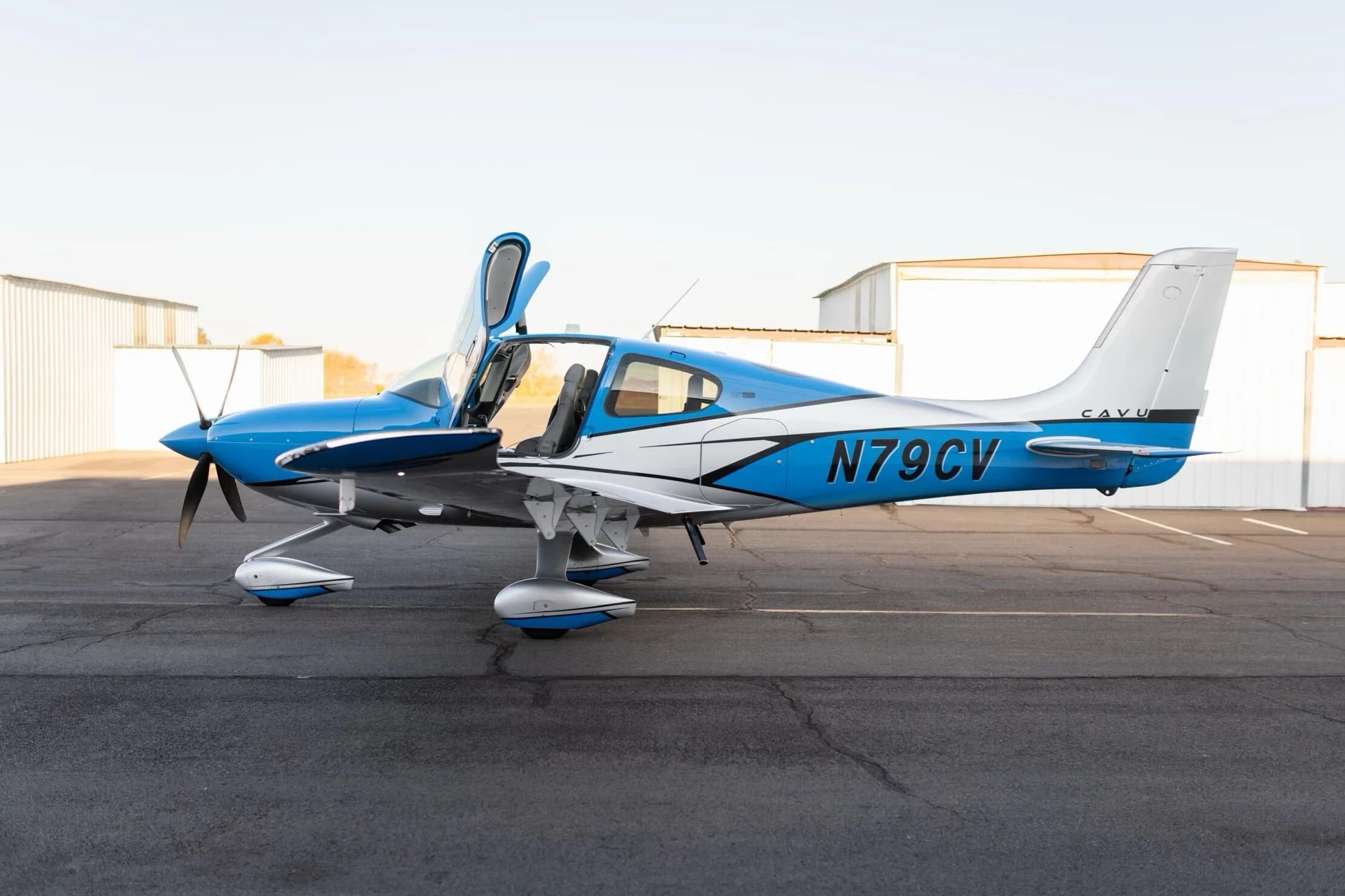 A side view of the blue and silver Cirrus SR22T GTS aircraft, registration N79CV, with its gull-wing doors open, parked on an airfield.