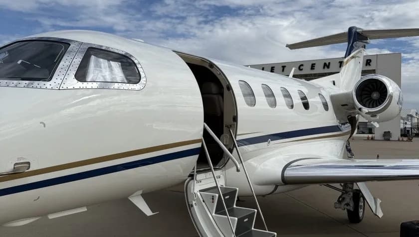 A close-up side view of a white Embraer Phenom 300 private jet with its passenger door open and stairs extended, parked at a jet center.