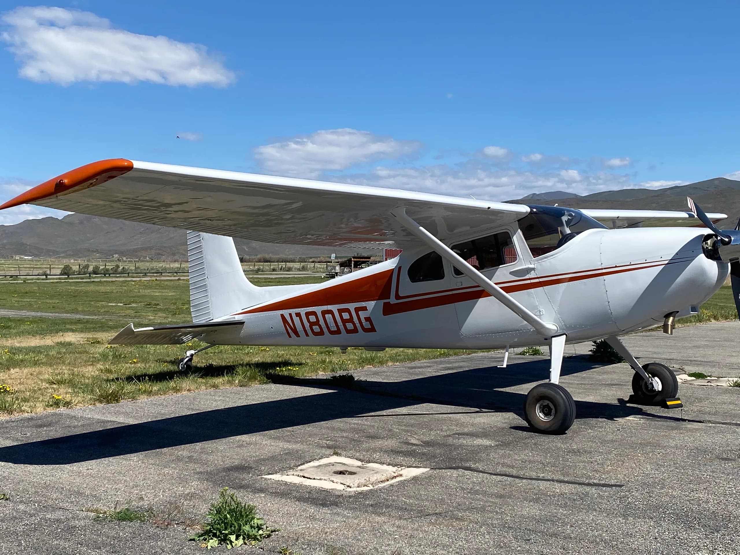 A white Cessna 170B with orange stripes and N180BG registration parked on an airfield with mountains in the background.