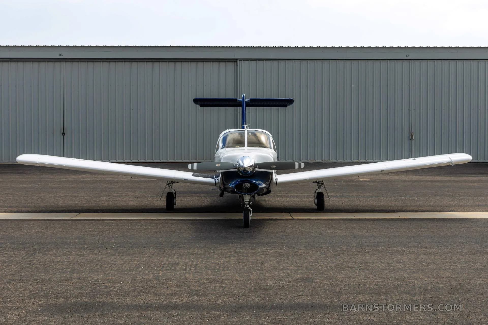 A front view of a white and blue single-engine propeller aircraft with a three-bladed propeller, parked in front of a hangar.