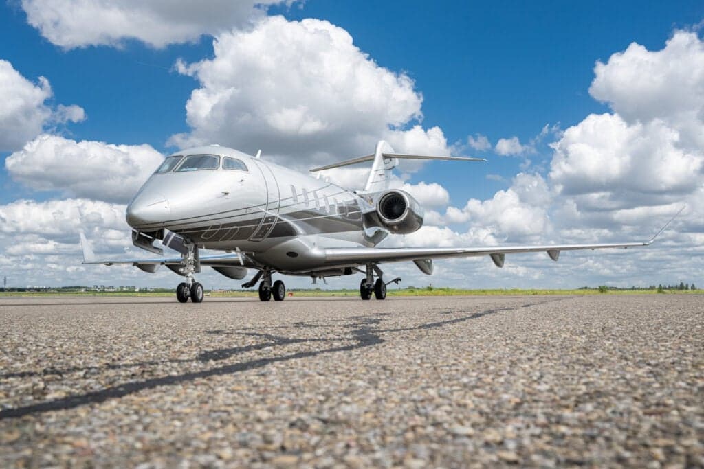 A silver and gray Challenger aircraft sits on the tarmac under a blue sky with clouds.