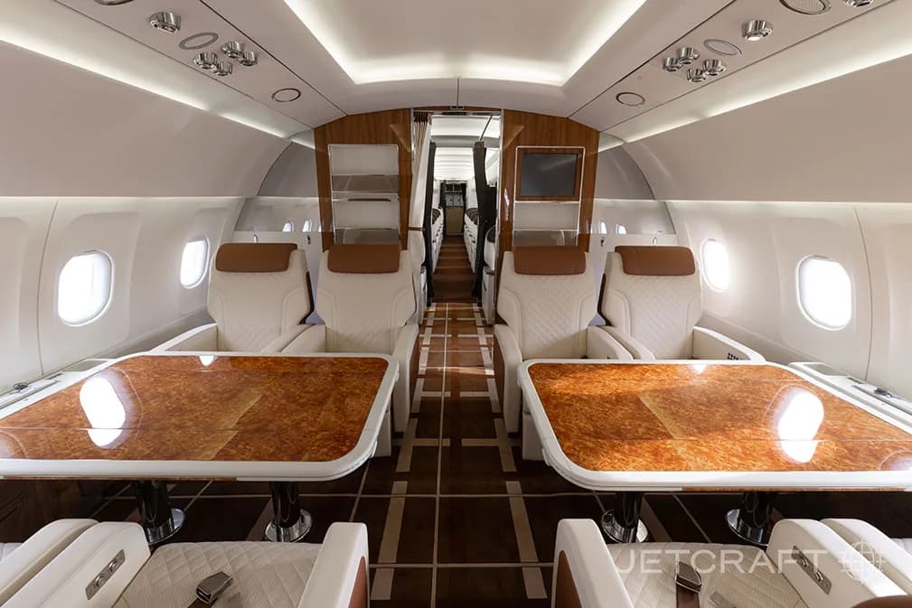 The luxurious interior of an aircraft cabin features two sets of facing seats with large polished wooden tables and light-colored upholstery.
