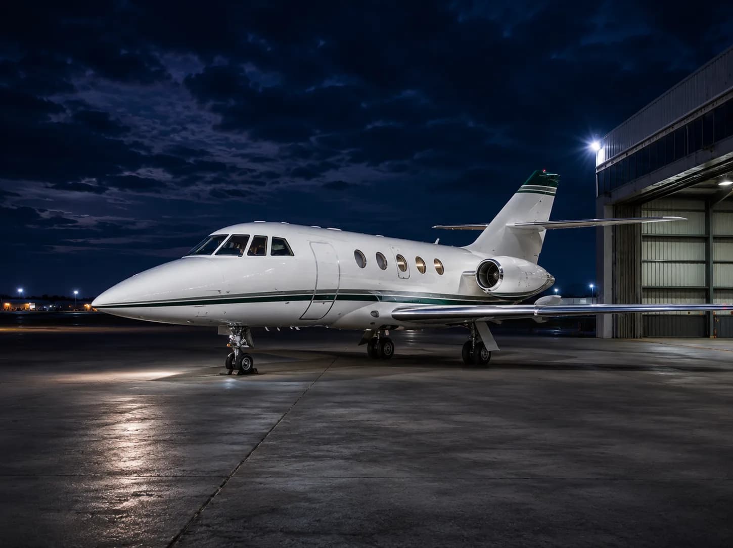 A white business jet with a green stripe is parked on a tarmac at night, illuminated by ground lights, with a dark cloudy sky.