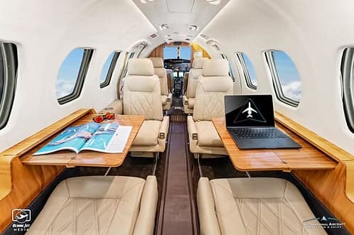The luxurious cabin interior of a business jet features light-colored leather seats, wooden tables, and a laptop.