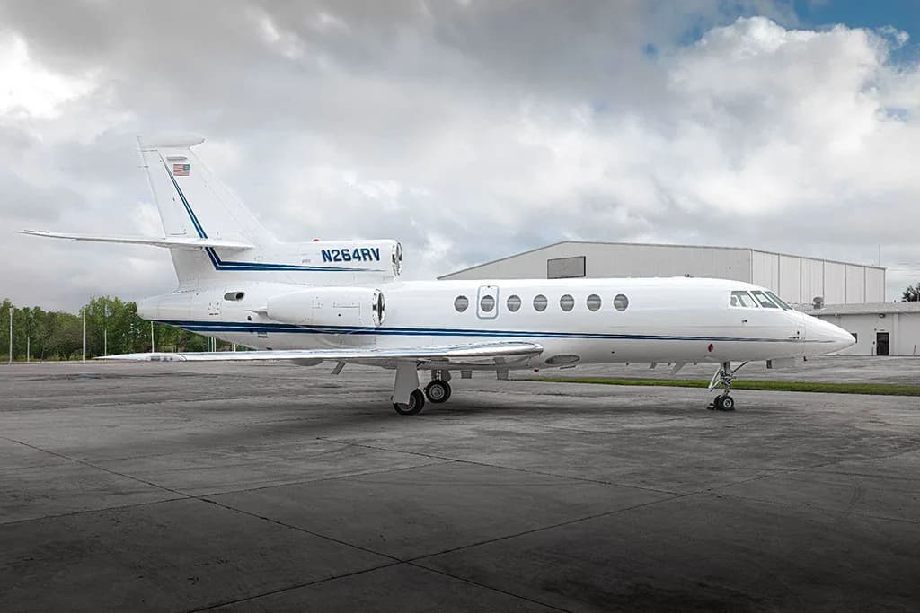 A white business jet with blue stripes, tail number N264RV, parked on an airfield under a cloudy sky.