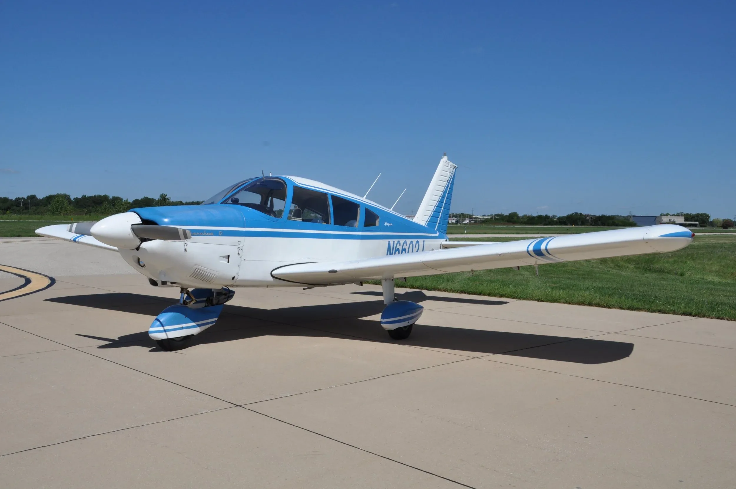 A white and blue Piper Cherokee D aircraft with registration N6600J is parked on an airfield under a clear blue sky.