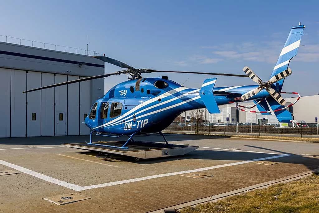 A rear-side view of a blue helicopter with white stripes, registration OM-TIP, on a helipad.