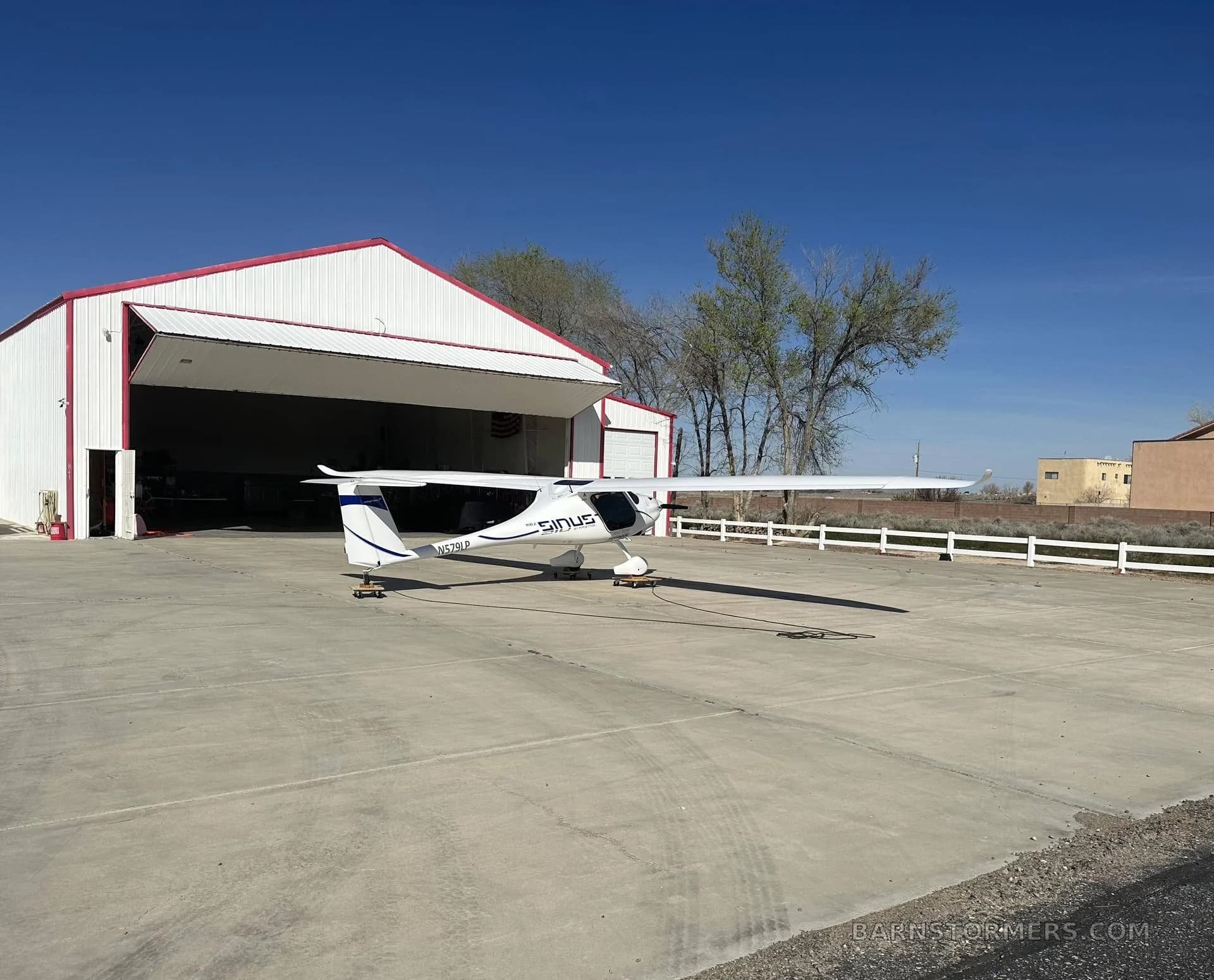 A white Pipistrel Sinus aircraft with blue accents is viewed from the rear-right, parked on an apron in front of a hangar.