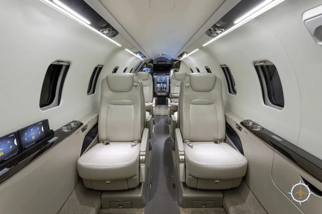 A luxurious aircraft cabin with light-colored leather seats and screens on the side panels, looking towards the cockpit.