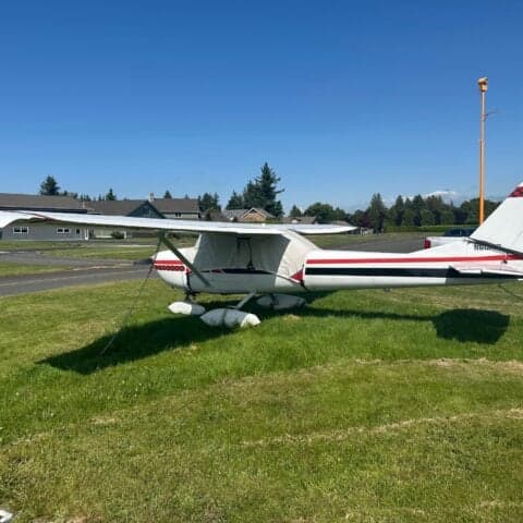 A Cessna 150 aircraft is parked on the grass with a white and black paint scheme and red accents.