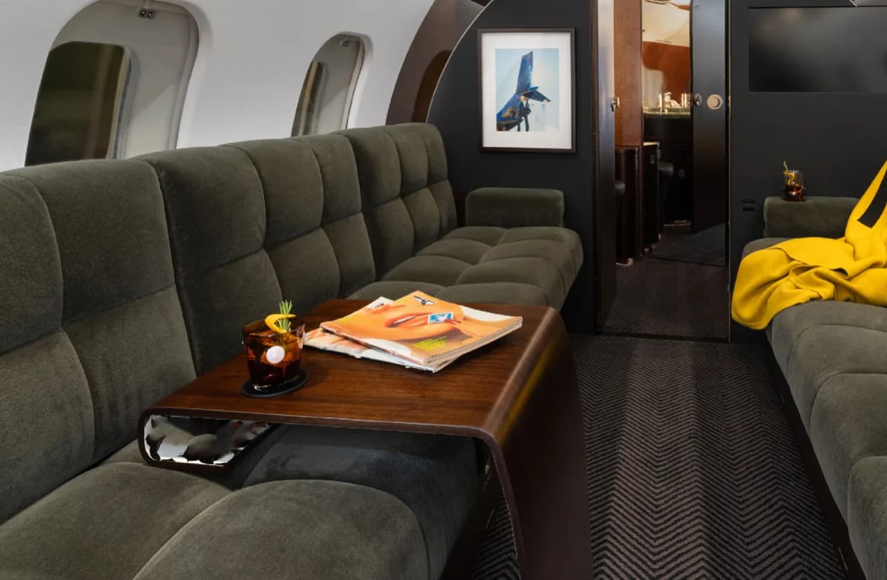 A comfortable lounge area in the private jet's cabin, featuring a dark green sofa, a wooden table with a drink and magazine, and a framed picture on the wall.