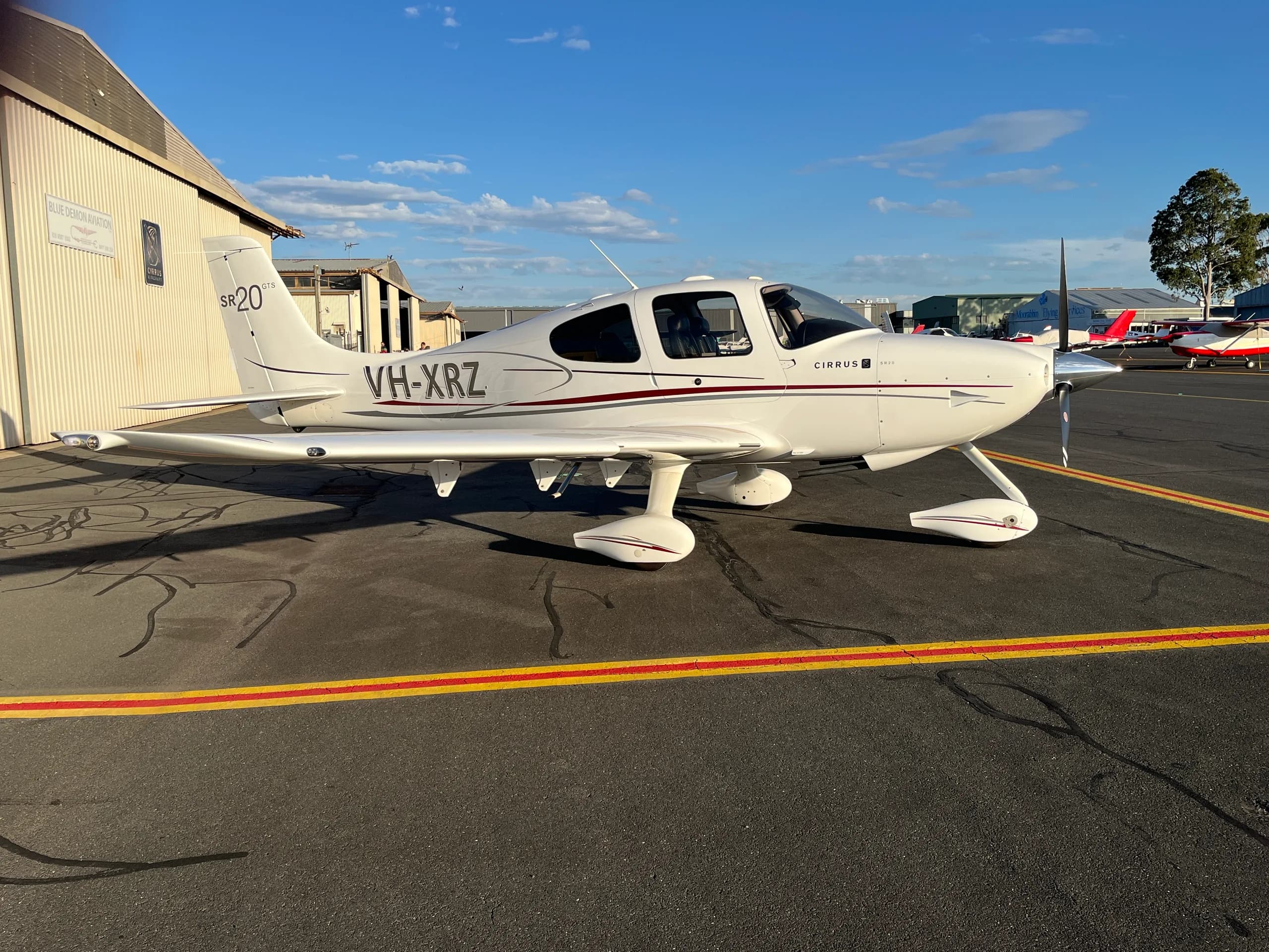 A white Cirrus SR20 GTS aircraft with red and grey stripes, registration VH-XRZ, parked on an airport tarmac next to a hangar.