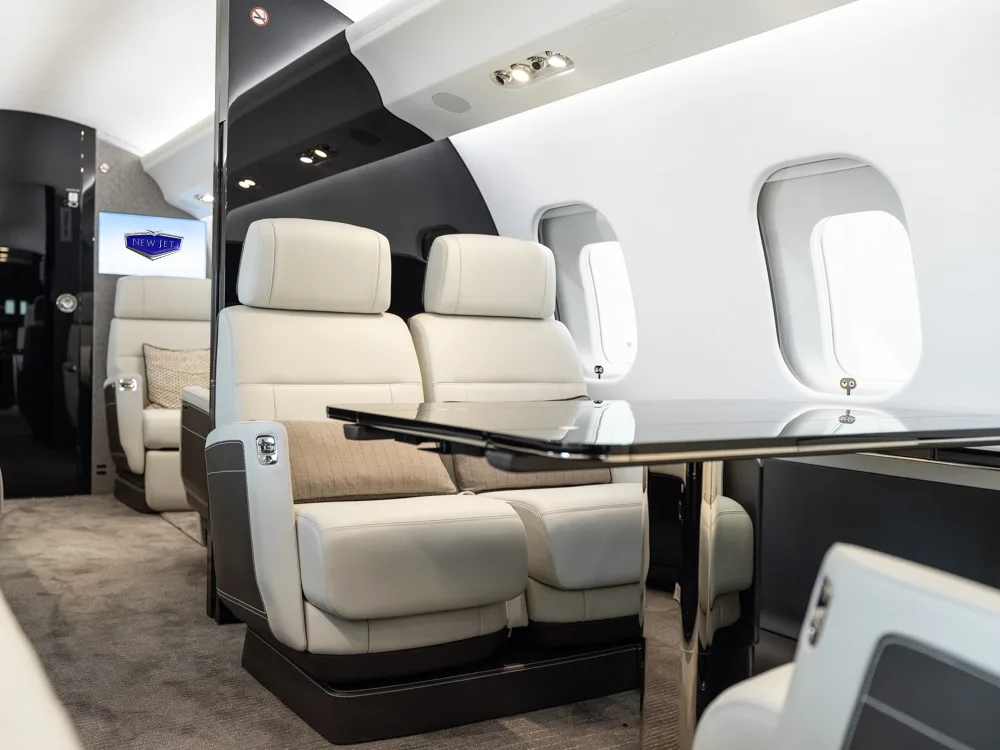 A view inside a private jet cabin showing two light grey seats facing a table, with a screen displaying a logo in the background.