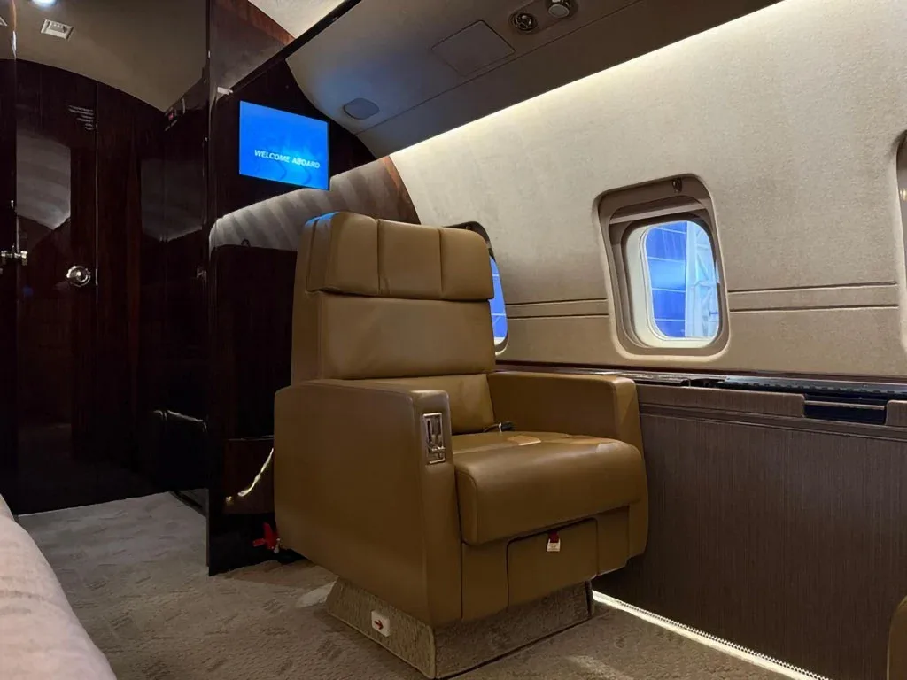 A tan leather seat in the private jet cabin faces a screen displaying "WELCOME ABOARD" and dark wood paneling.
