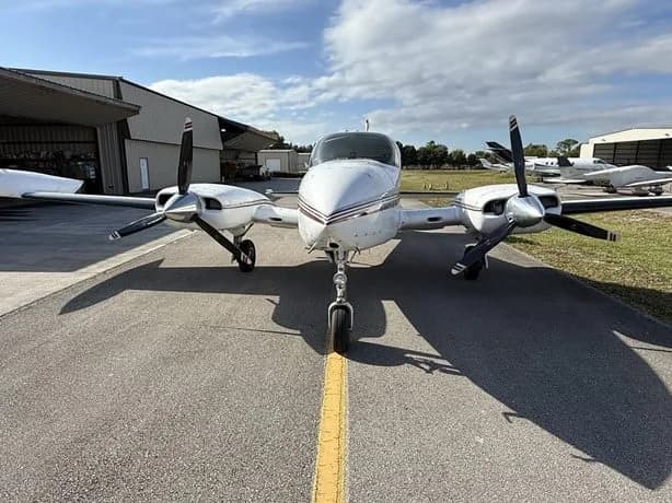 A white twin-engine aircraft with red and black stripes, viewed from the front on an airfield.