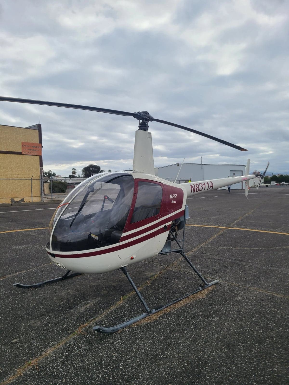 A white and red Robinson R22 Beta helicopter, registration N8311A, parked on an airport apron under a cloudy sky.