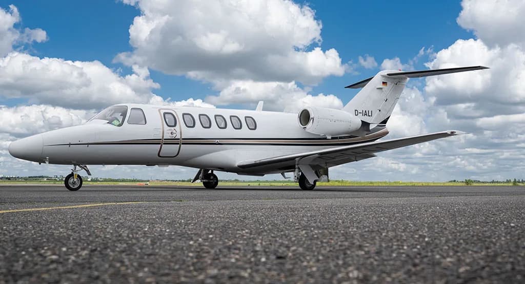 A white Cessna Citation business jet with black and brown stripes, registration D-IALI, parked on an asphalt runway under a blue sky with clouds.