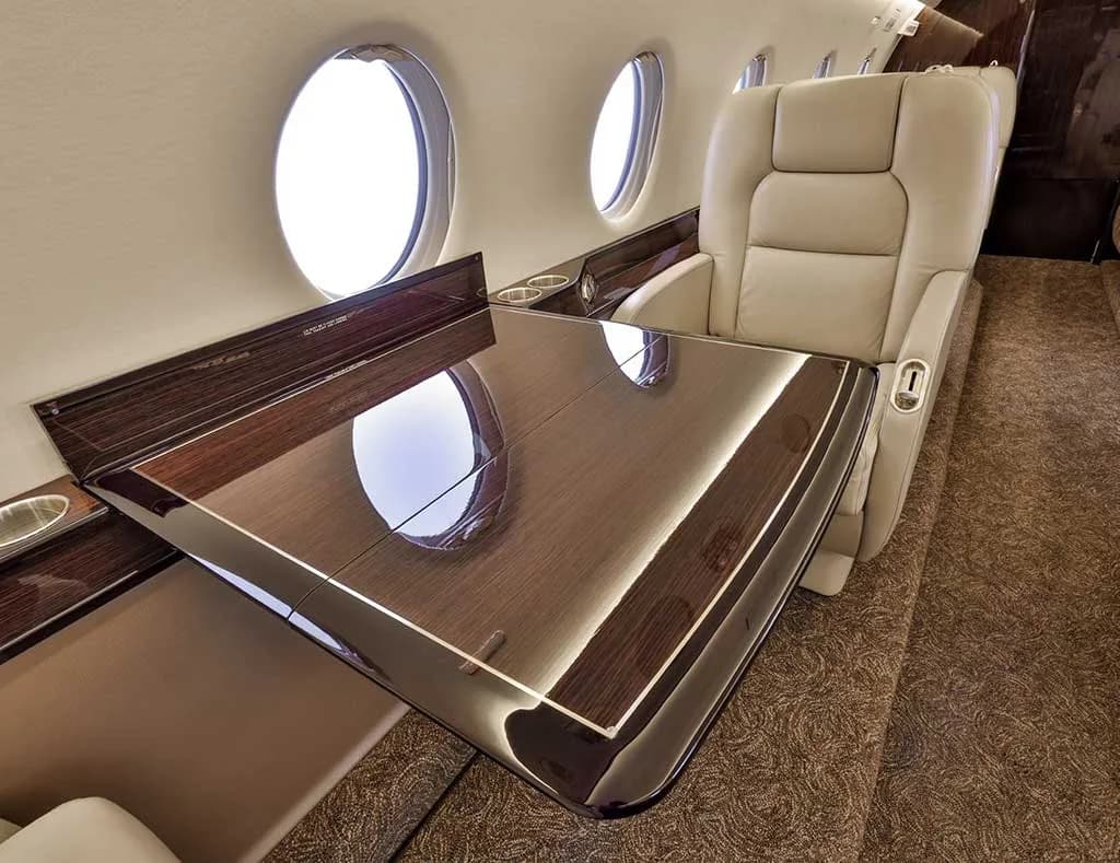A polished wooden table extended in the private jet's cabin, next to a beige leather seat and a window.