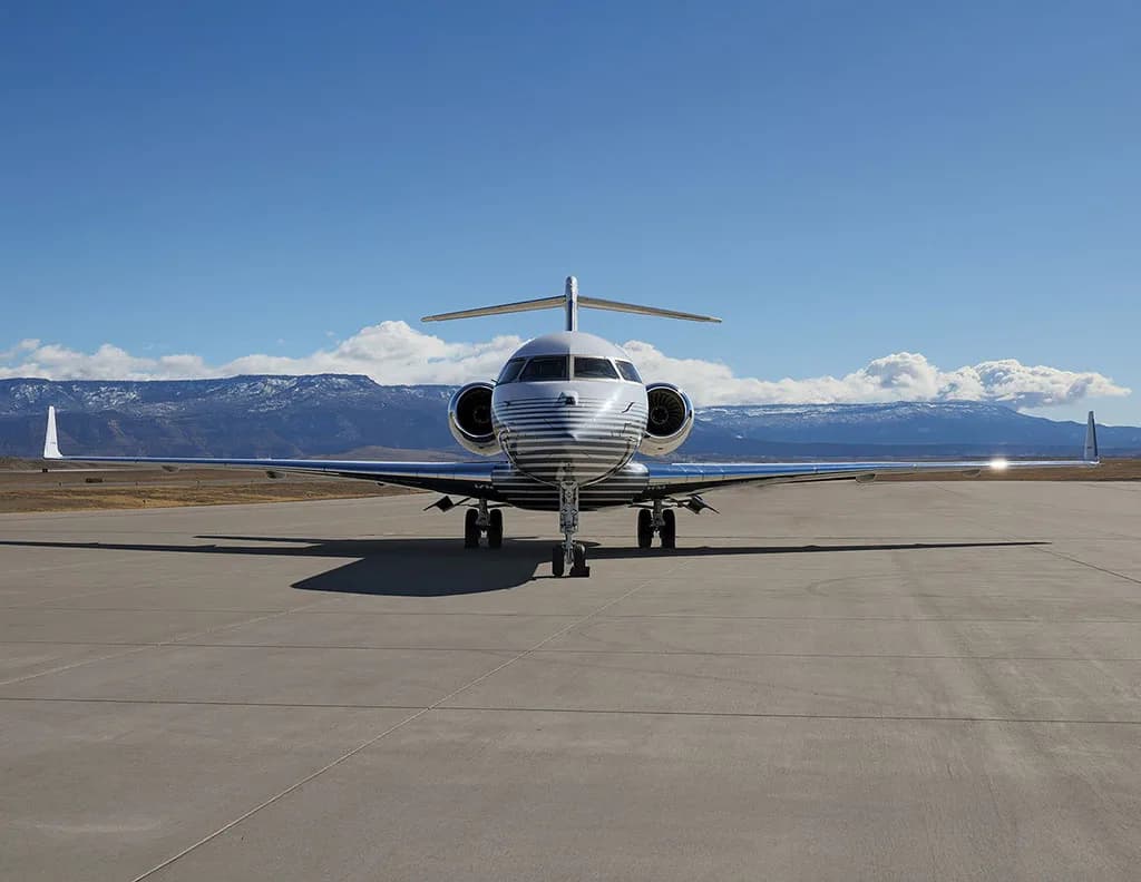 A head-on view of a white business jet with grey stripes on the fuselage, parked on an airfield with mountains in the distance.