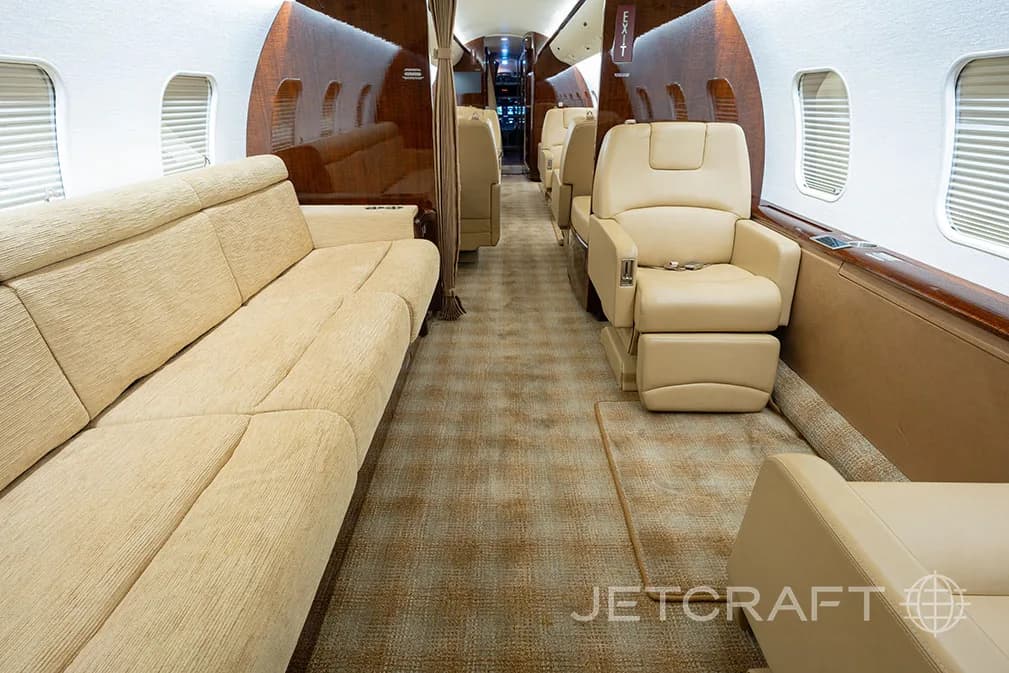 The luxurious interior cabin of a private jet featuring a beige sofa, individual seats, and wood paneling.