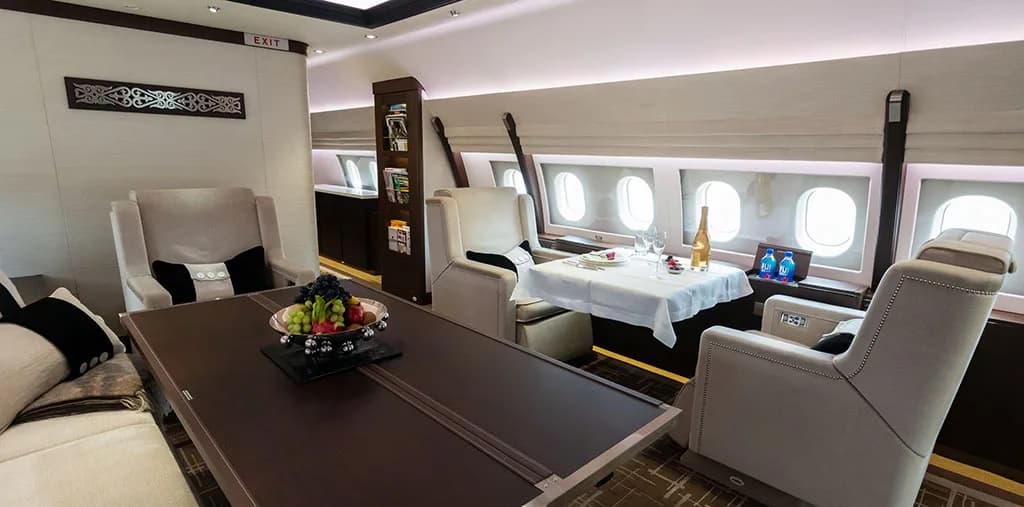 A spacious aircraft cabin featuring a dining table set for two, lounge chairs, and multiple windows.