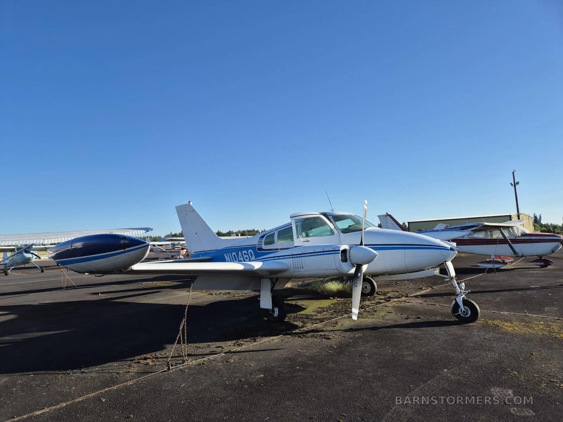A white twin-engine aircraft with blue stripes and registration N1046Q, seen from the front-left, parked on an asphalt ramp with other aircraft in the background.