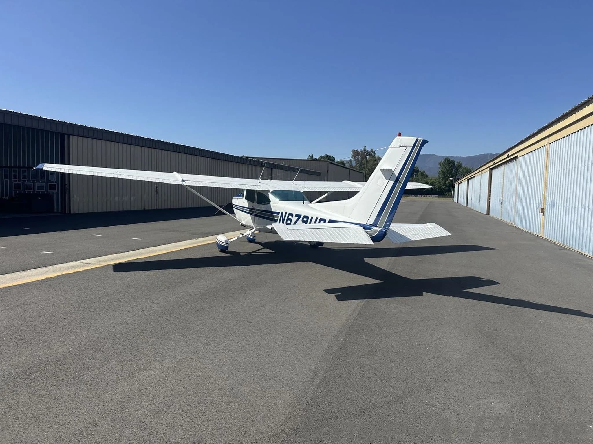 A white and blue Cessna 182 Skylane aircraft parked on an asphalt ramp, viewed from the rear-left.