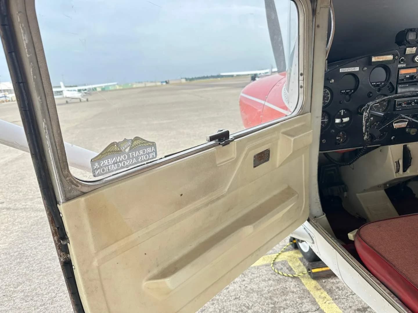 View from outside the open door of the Cessna 150, showing part of the instrument panel and an airfield outside.