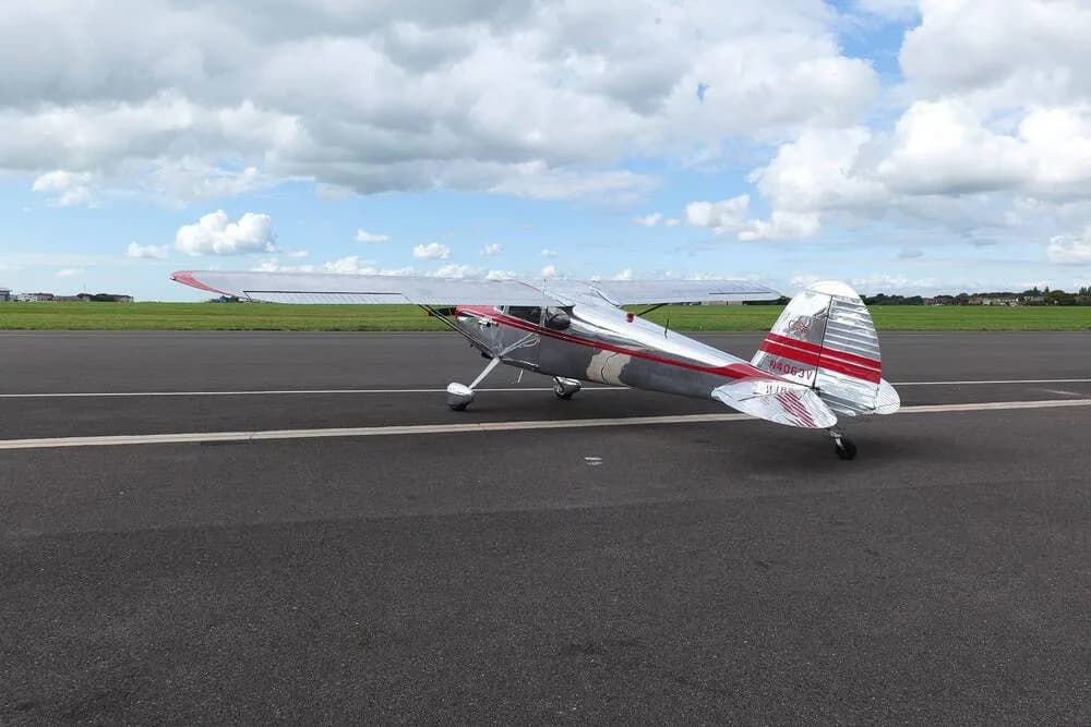 A three-quarter rear view of a shiny silver Cessna 170B with red stripes, parked on an empty tarmac.