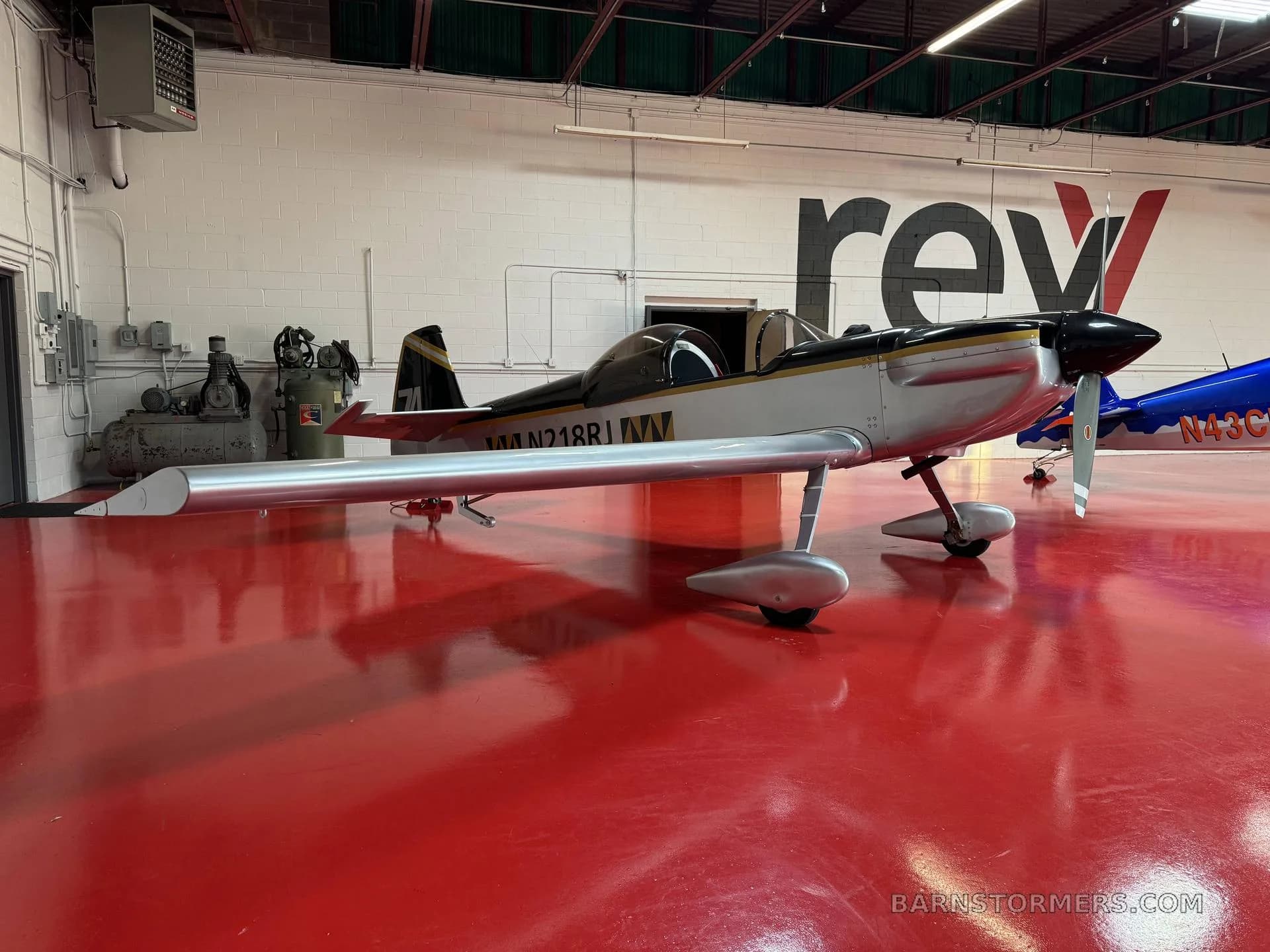 A silver and black single-engine aircraft with registration N218R is parked on a red floor in a hangar.