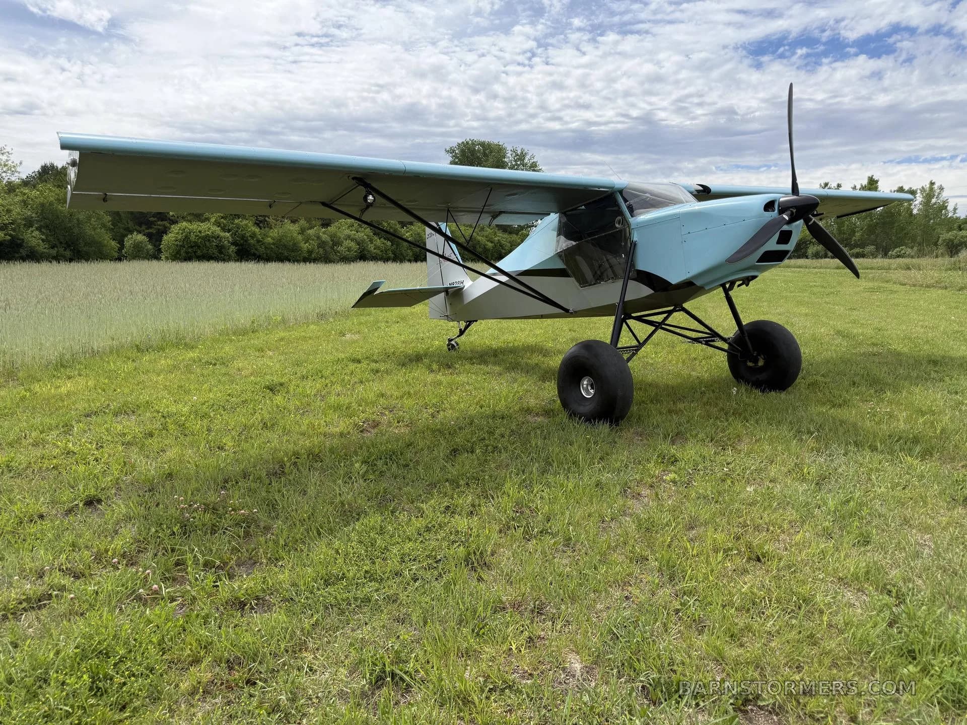 A front-right view of a light blue and grey Just Aircraft SuperSTOL with large tundra tires, parked on a grassy field.
