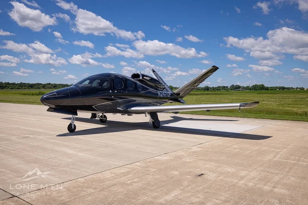 A black Cirrus Vision Jet (N247CF) with silver wings and accents parked on a tarmac next to a grassy field.