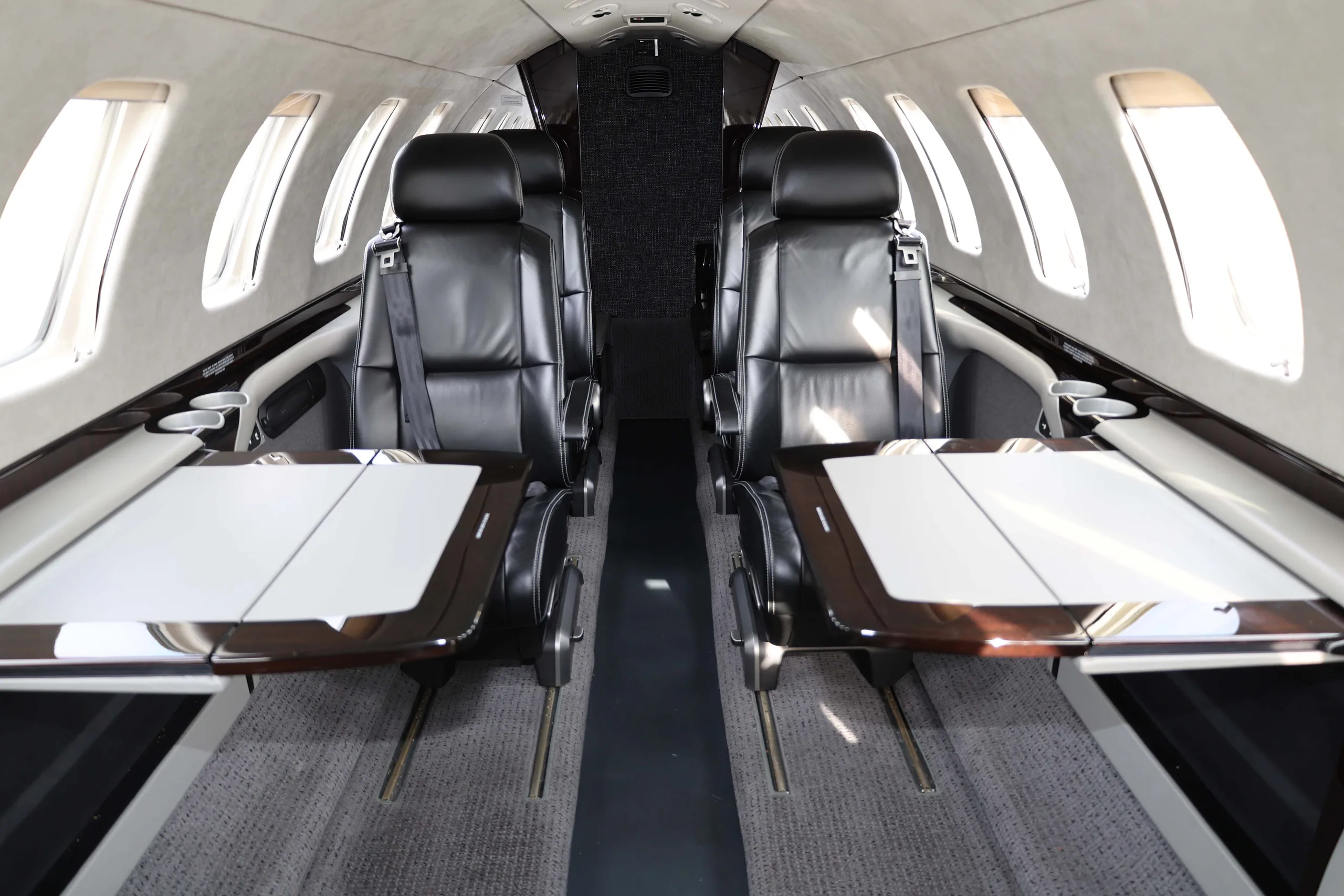The interior cabin of a business jet, featuring black leather seats and two fold-out tables with dark wood and white surfaces.