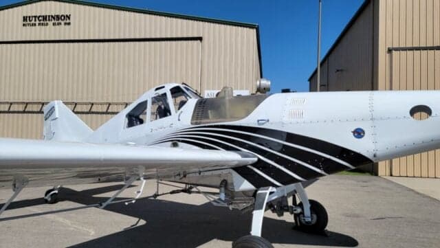Side view of a white agricultural aircraft with black stripes parked on an tarmac in front of a hangar.