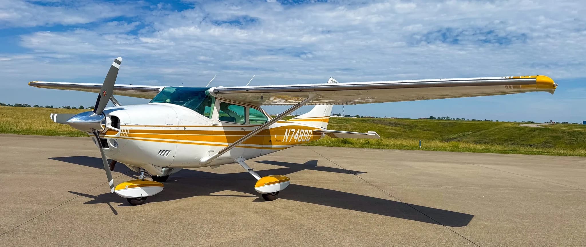 A white and gold Cessna Skylane N7489Q is parked on an airfield tarmac under a partly cloudy sky, viewed from the front-left.