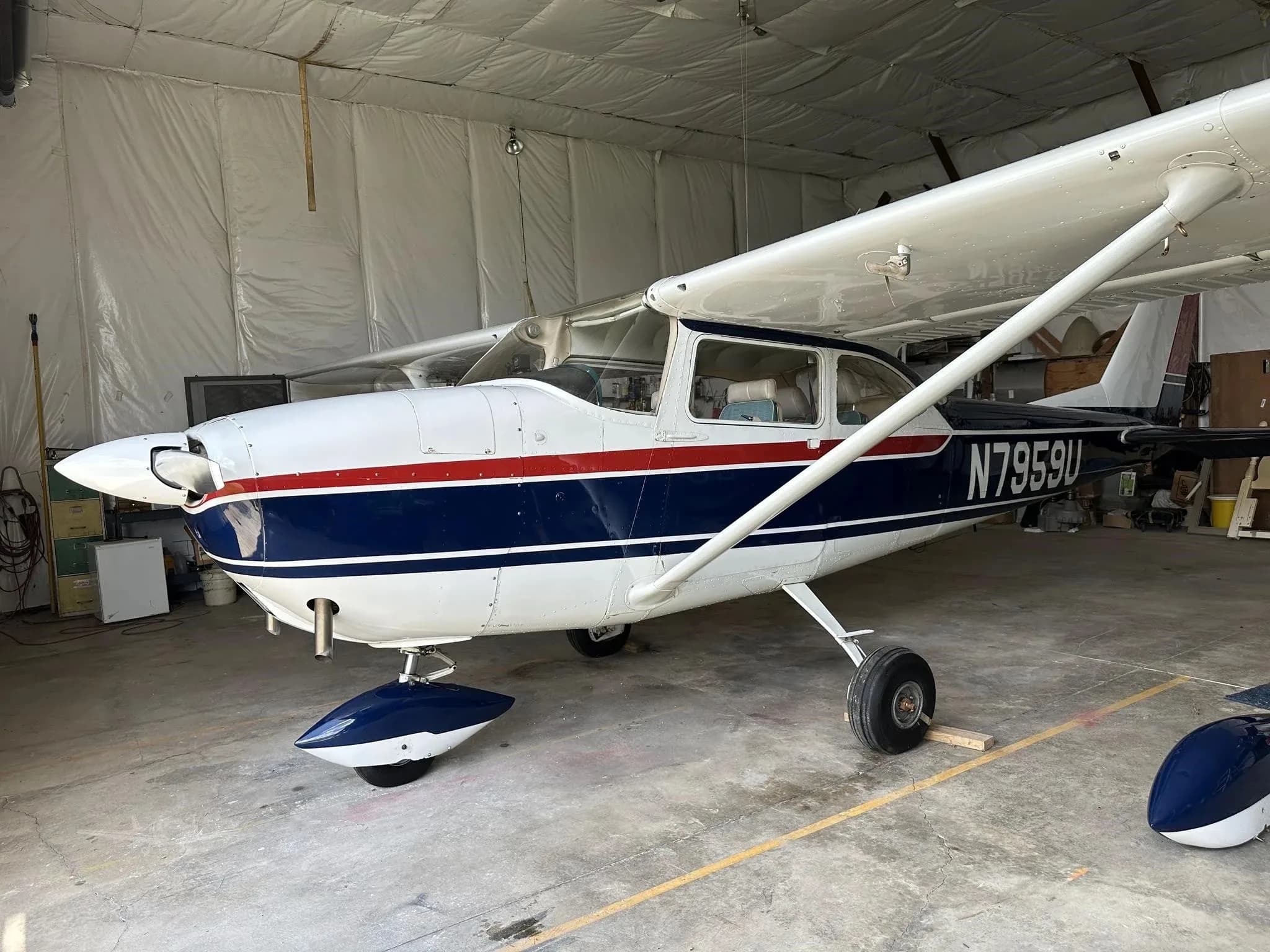 A side view of the white and blue Cessna N7959U with a red stripe in a hangar, with a wheel fairing removed.