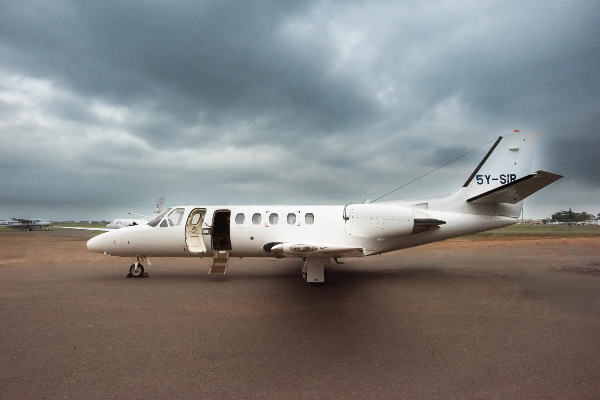 A white Cessna Citation business jet with registration 5Y-SIR parked on a tarmac with its main door open under a cloudy sky.