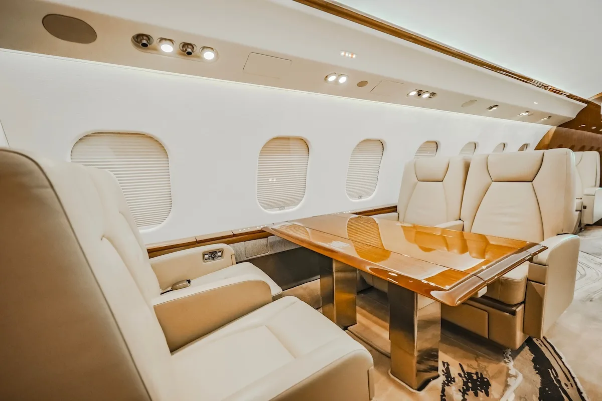 A private jet cabin interior with light-colored seats arranged around a large wooden table.