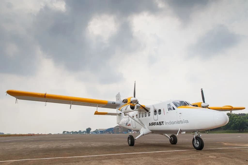 A white and yellow Airfast Indonesia Twin Otter aircraft parked on an airfield under a cloudy sky.