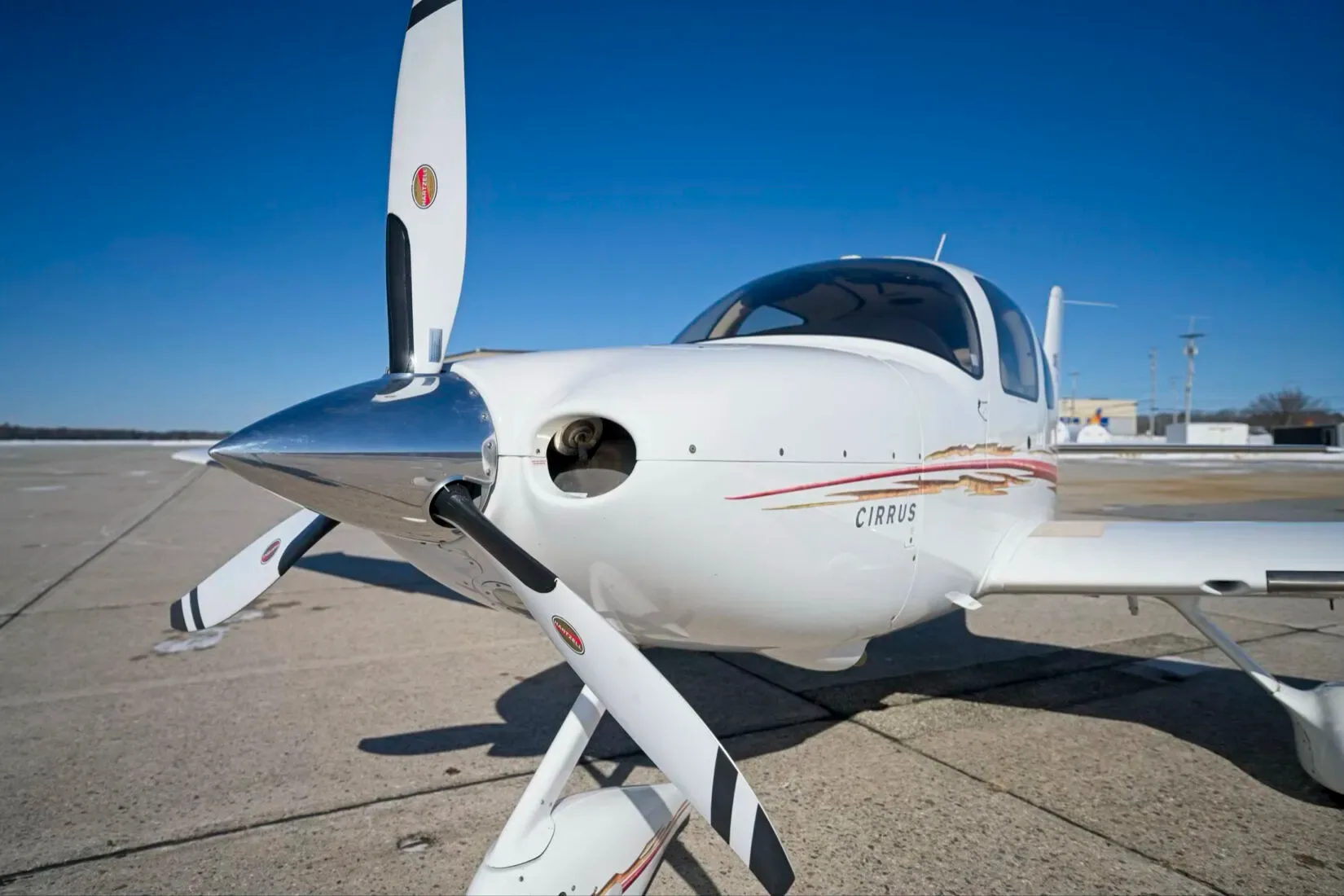 A close-up front view of a white Cirrus SR22 aircraft with a polished spinner and red and gold stripes.