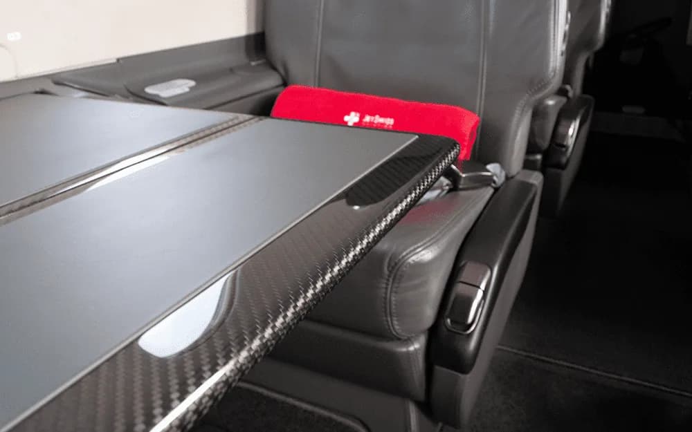 A close-up view of a dark grey leather aircraft seat with a red pillow and a carbon fiber-trimmed fold-out table.