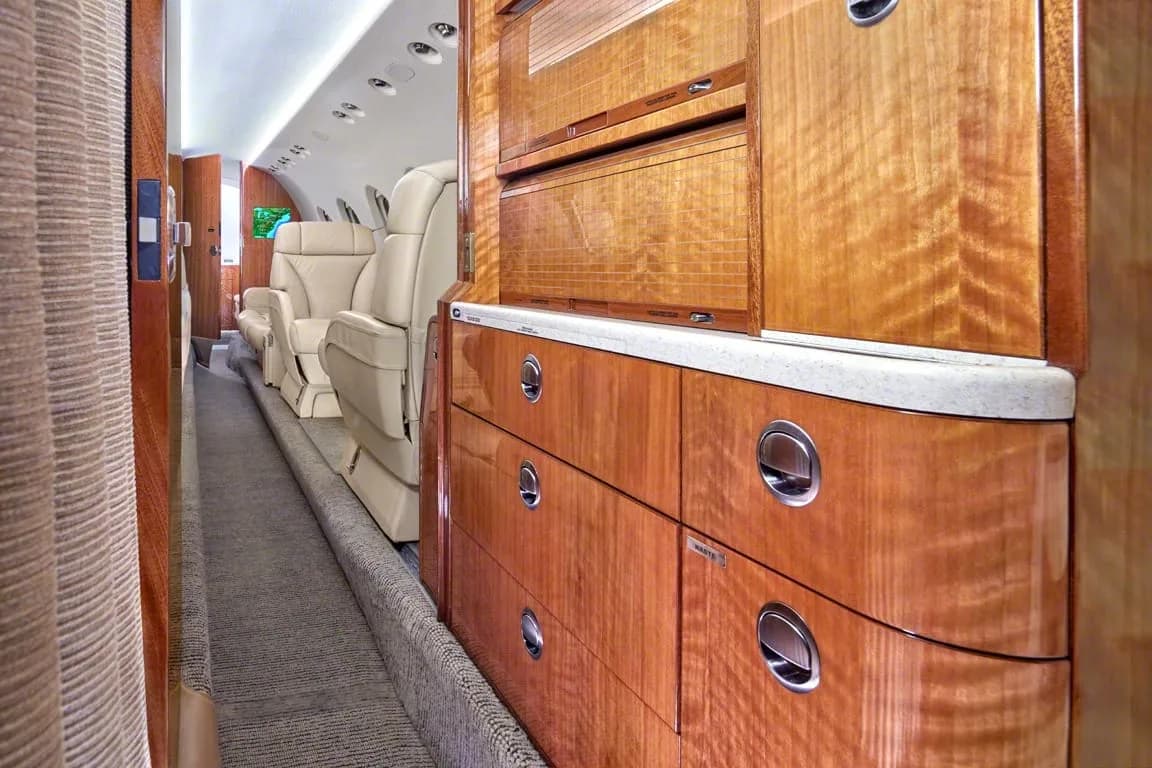 A view down the aisle of a private jet cabin, highlighting polished wood cabinetry and beige seats.