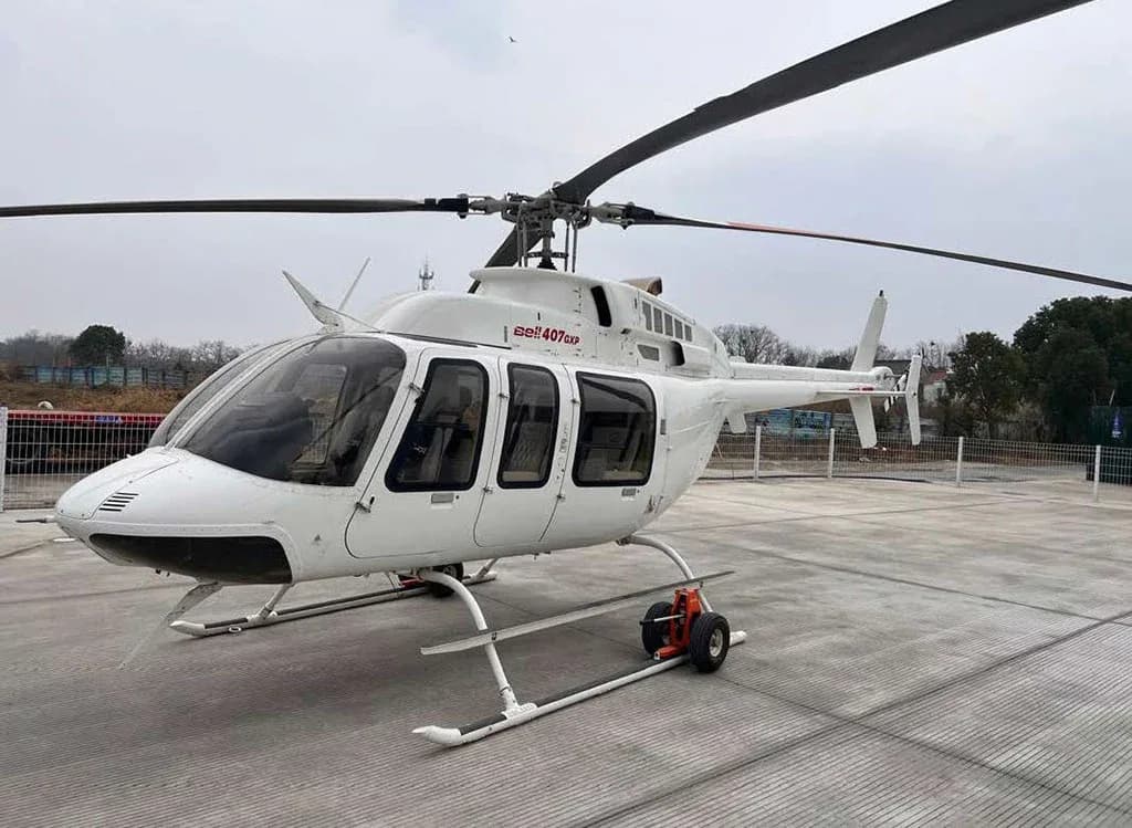 A white Bell 407GXP helicopter with landing skids and ground handling wheels, viewed from the front-left on a concrete pad.