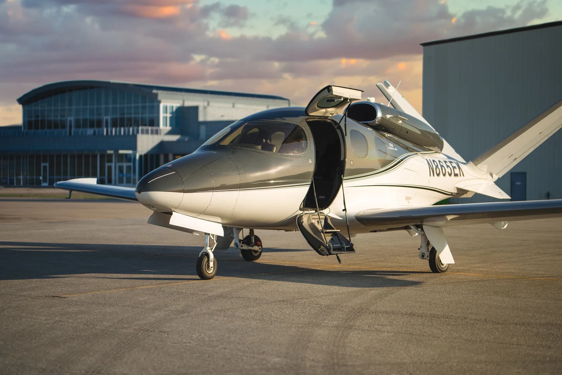 A Cirrus Vision Jet (N865EK) with its door open is parked on an airfield in front of a modern hangar.