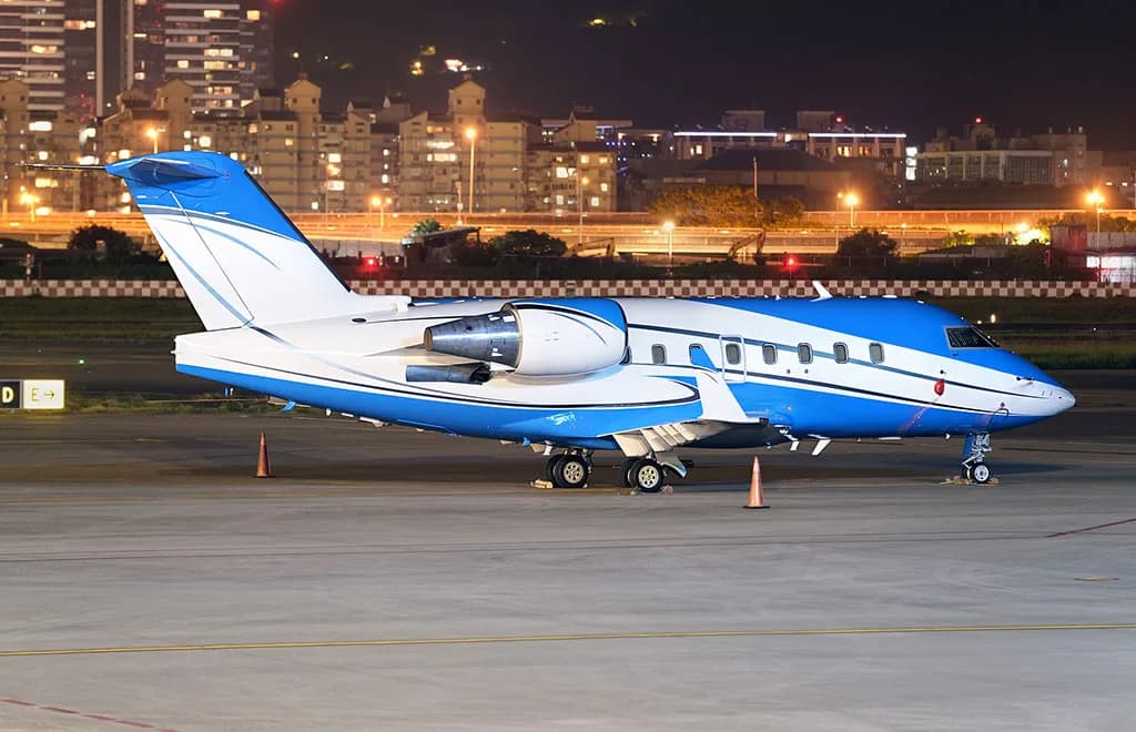 A blue and white Bombardier Challenger 604 business jet is parked on an airport tarmac at night, with a city skyline illuminated in the background.